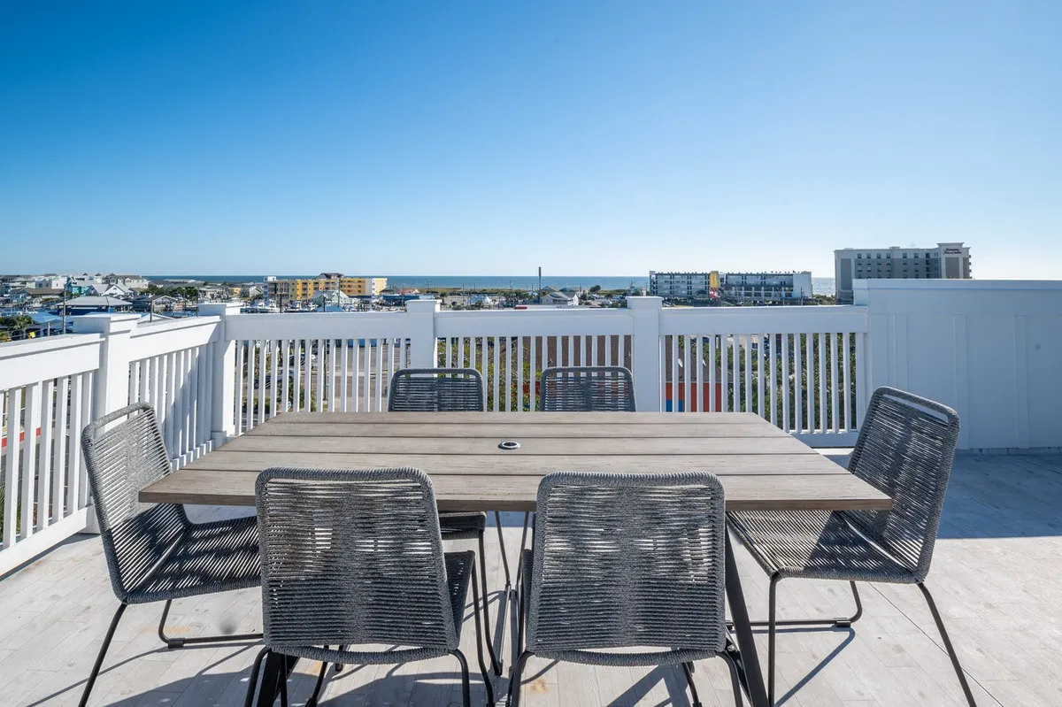 The image showcases a spacious rooftop deck with a dining table set for a gathering, offering sweeping views of the ocean and surrounding Carolina Beach boardwalk. The atmosphere is relaxed and inviting, perfect for outdoor dining, sunset watching, and celebrating with friends or family.
