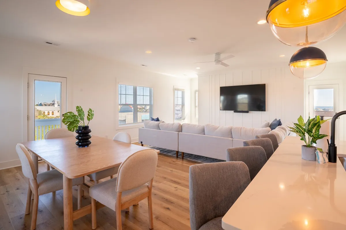 This image showcases an inviting open-plan living area with a large sectional sofa, dining table, kitchen island, modern lighting fixtures, and a flat-screen TV. The space is filled with natural light from expansive windows, creating a bright, cheerful atmosphere ideal for family gatherings or relaxing after a day at the beach.