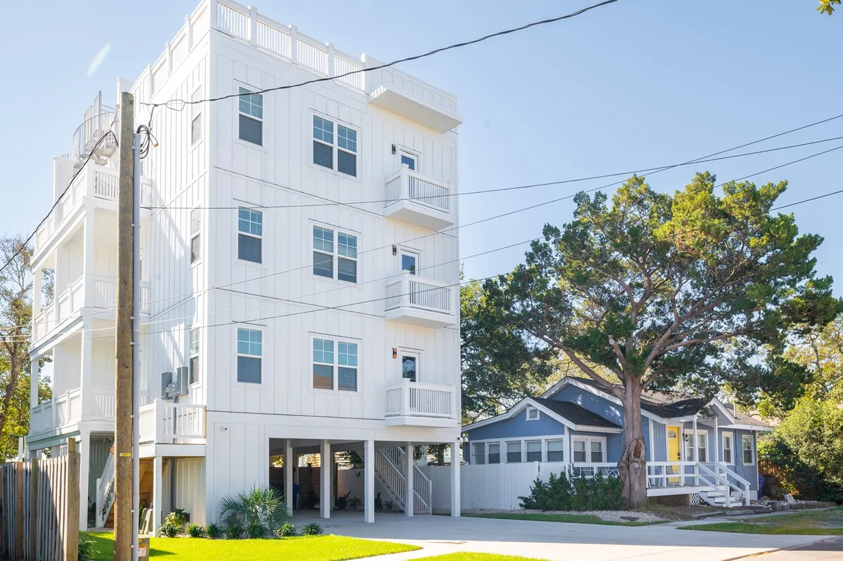 A cheerful exterior shot showing a modern multi-story duplex next to a traditional house, with a large tree and green lawn in the foreground. The balconies offer glimpses of sunset views and spaces for outdoor gatherings, while the fenced yard emphasizes comfort and family-friendly amenities.