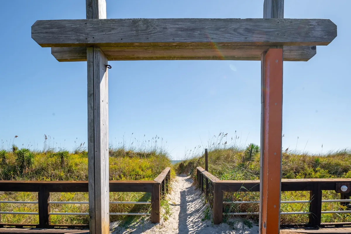 This image features a tranquil wooden archway framing a pathway that leads directly to the sandy beach, surrounded by lush greenery. The inviting arch signals the transition from neighborhood to Carolina Beach's welcoming shores, reinforcing the ease of access highlighted in the section.