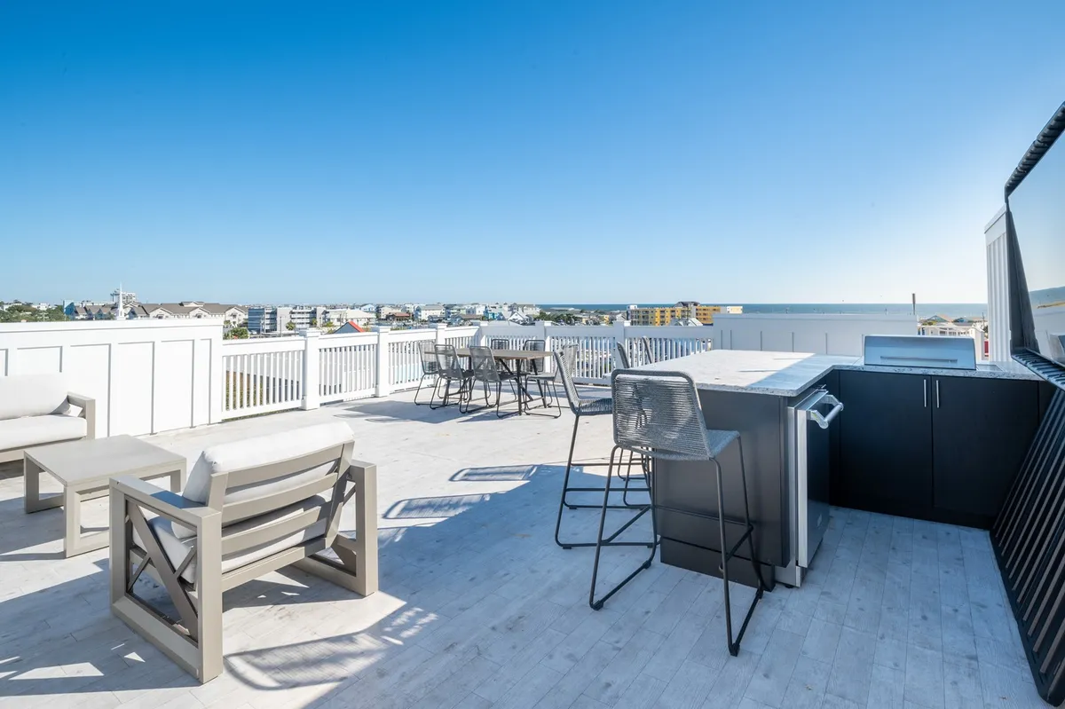 A spacious rooftop deck with an outdoor seating area, a barbecue grill, a dining table, and a sweeping ocean view. The scene is bathed in golden sunset light, evoking a sense of relaxed luxury and ideal conditions for hosting gatherings or quiet moments alike.