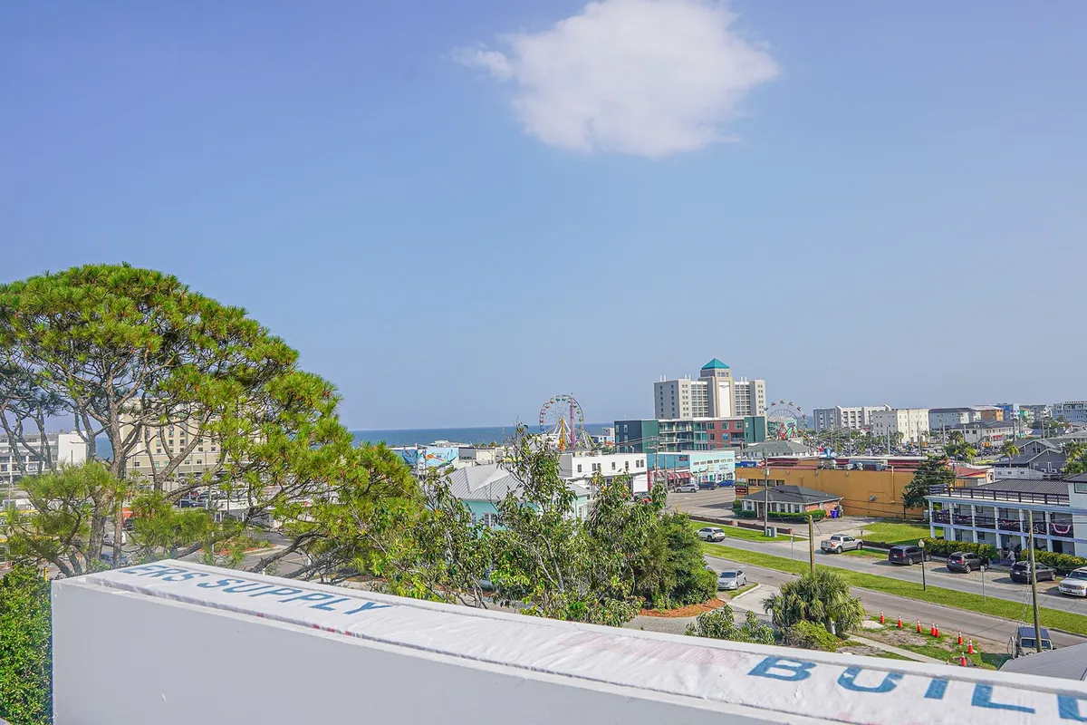 A vibrant view featuring the Ferris wheel and amusement park rides along the Carolina Beach Boardwalk, with the ocean, city buildings, and green trees in the background. The image captures the lively, playful atmosphere of the boardwalk area and the allure of classic seaside amusements.