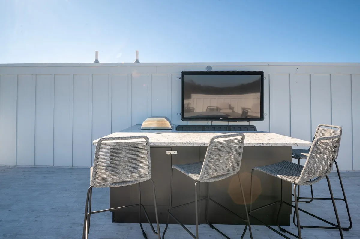 An exterior scene showing a stylish rooftop bar counter with tall stools and a mounted television, set against an open sky backdrop. The space is designed for outdoor dining, socializing, and enjoying sunset views with friends or family.