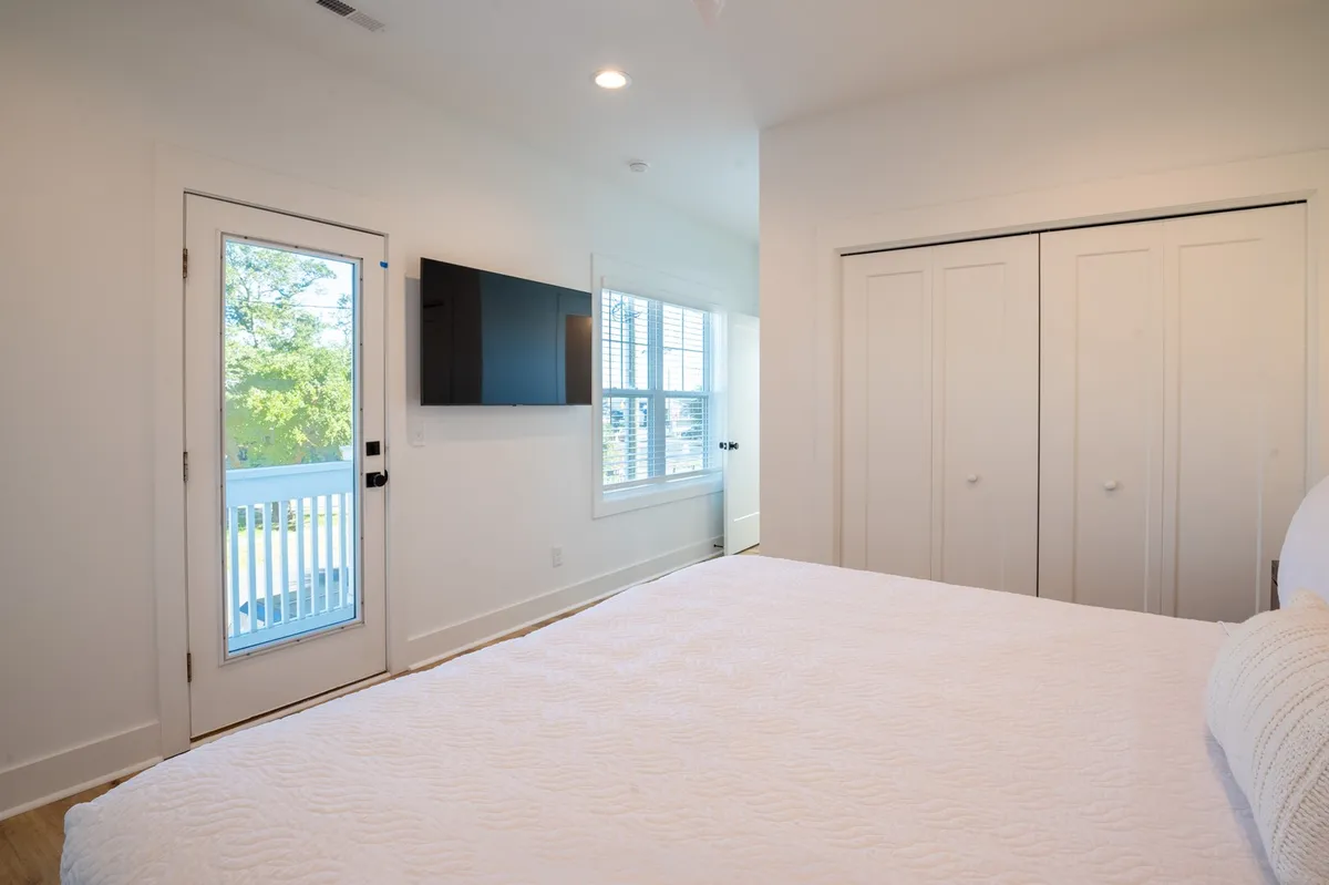 A serene king bedroom with white bedding, a wall-mounted TV, a door leading to a private balcony, and large windows letting in natural light. The inviting decor and peaceful ambiance suggest a restful, upscale retreat.